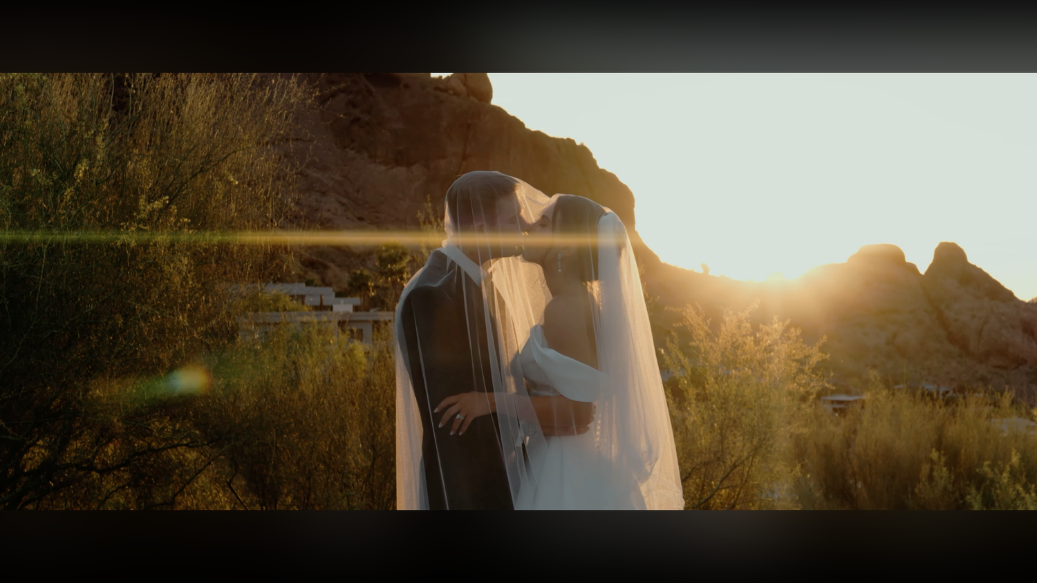 A bride and groom share a romantic kiss at sunset in a desert landscape with golden light and rocky mountains.