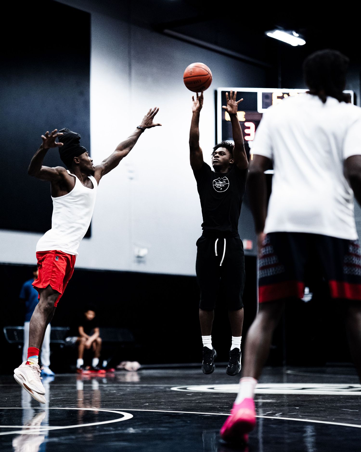 A basketball player in black shorts rises for a jump shot over a defender in an indoor gym.