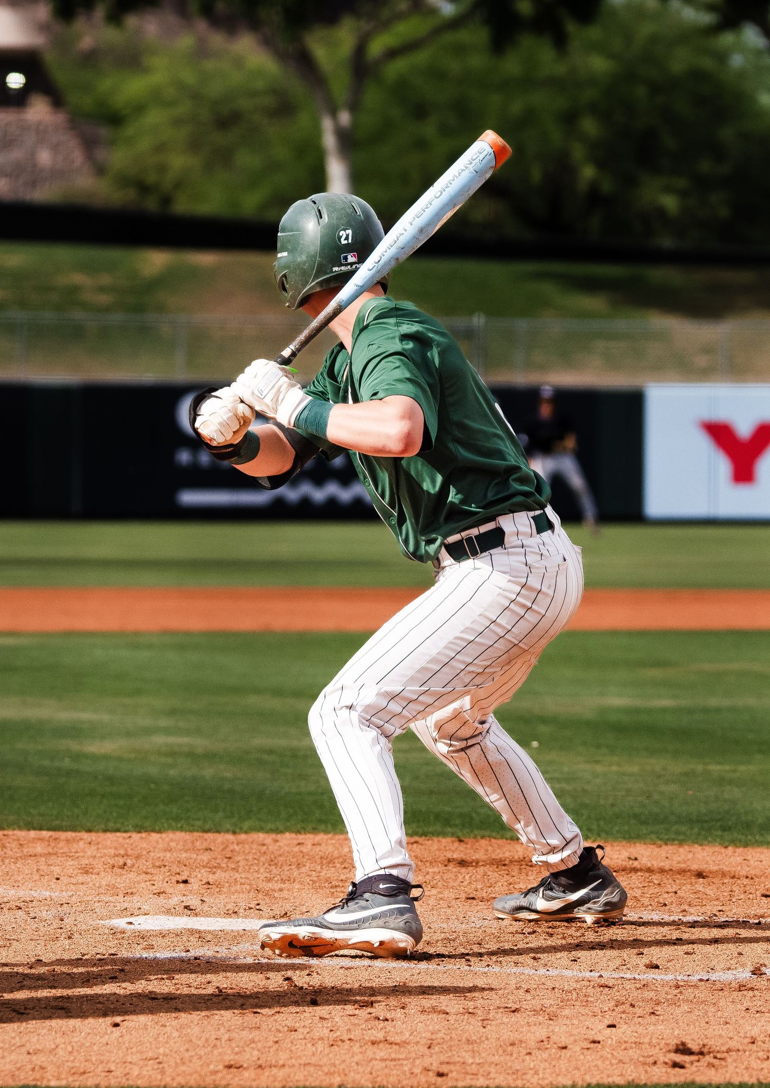 Baseball batter in green helmet and uniform prepares to swing at pitch during a sunny outdoor game.