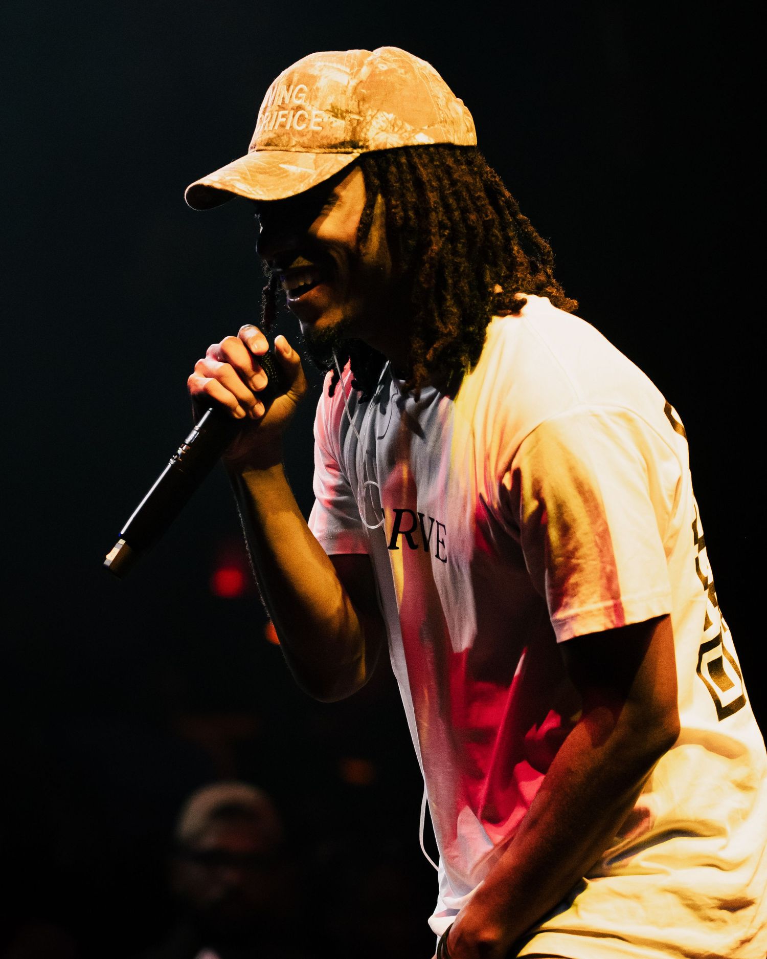 A performer with dreadlocks wearing a cap and white t-shirt singing into a microphone under stage lights.