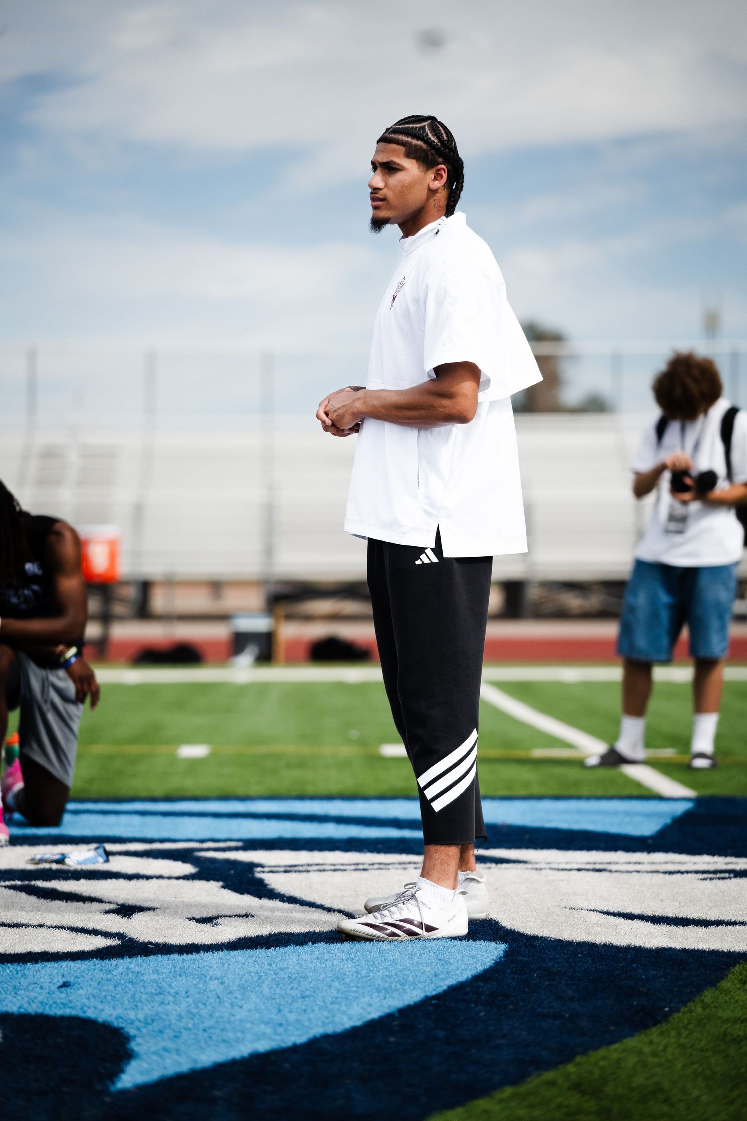 Young male athlete in white Adidas shirt and black pants stands on blue turf football field.