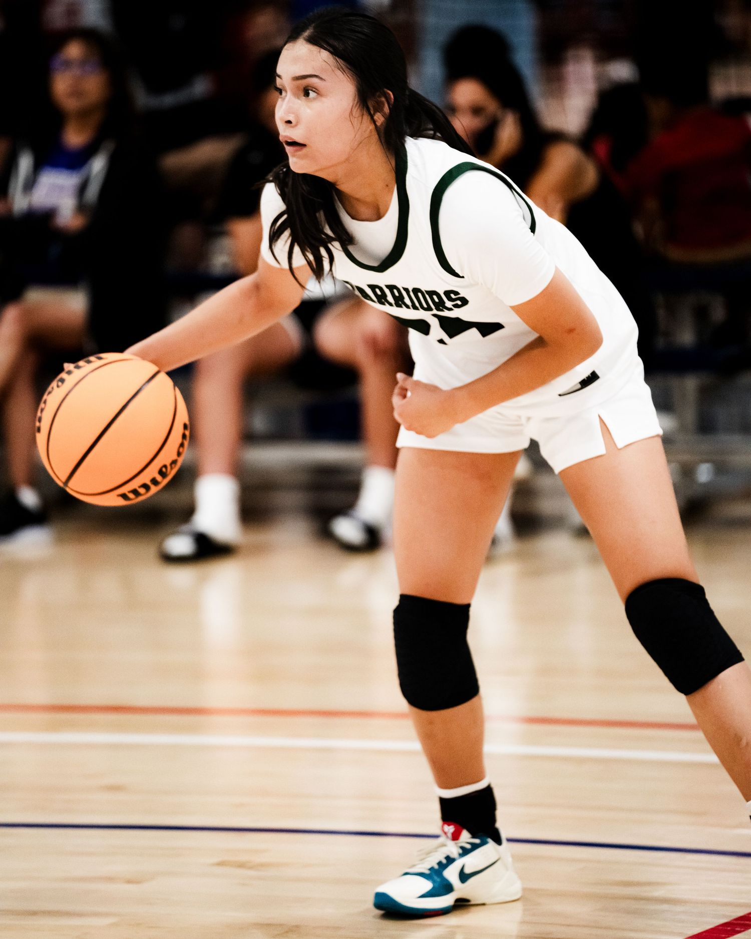 Female basketball player in white Warriors uniform dribbling ball on court wearing knee pads and Nike shoes.