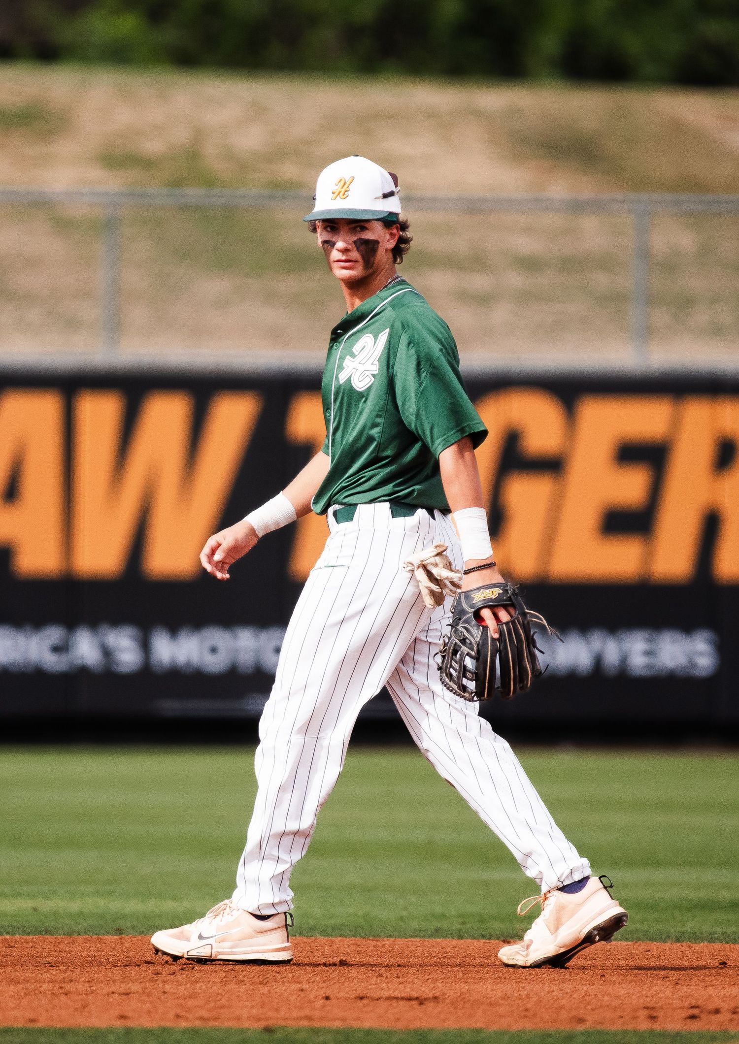 Baseball player in green uniform smiling on field near dugout signage.