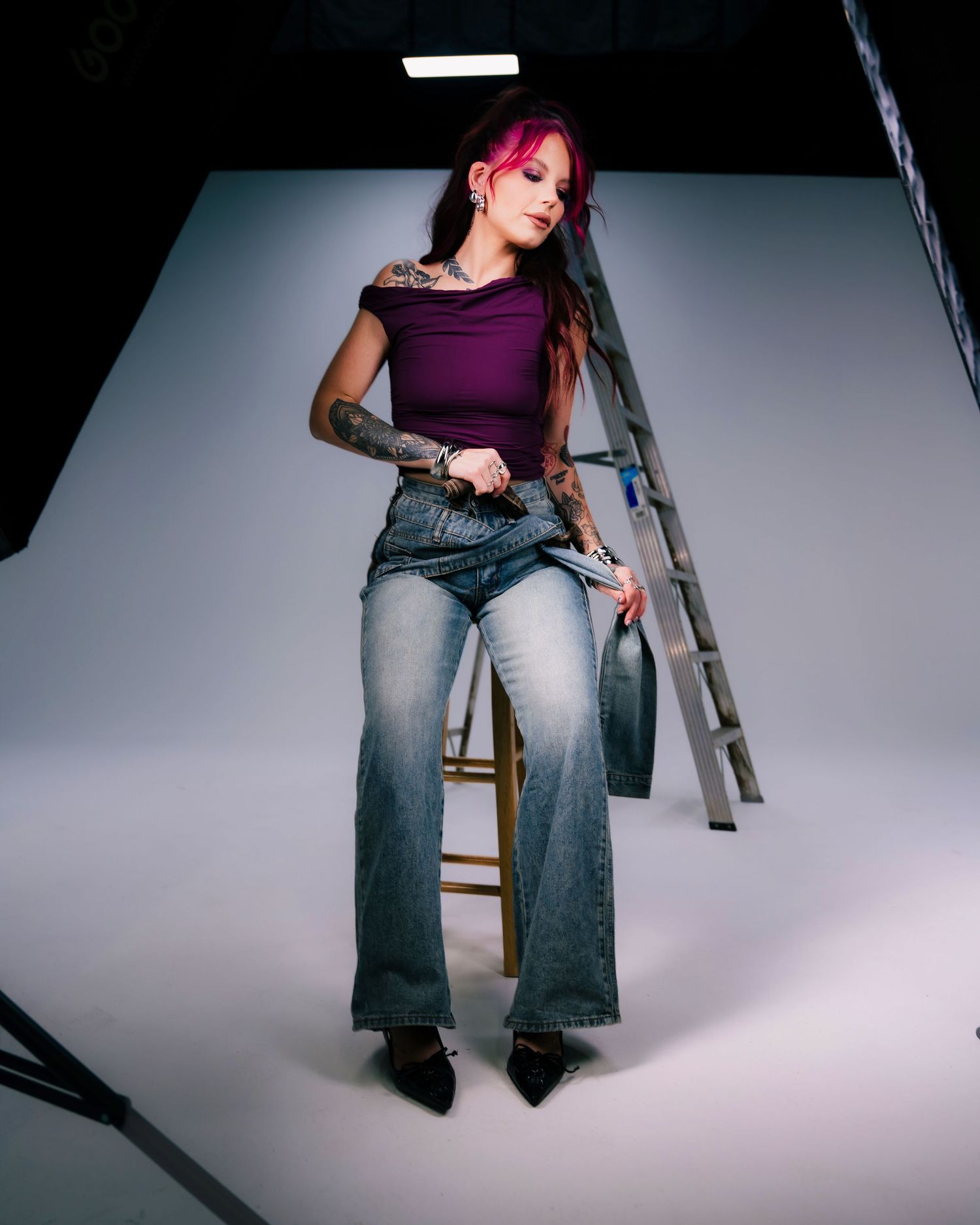 Tattooed woman with pink hair in purple crop top and wide-leg jeans poses on stool near ladder in photo studio.