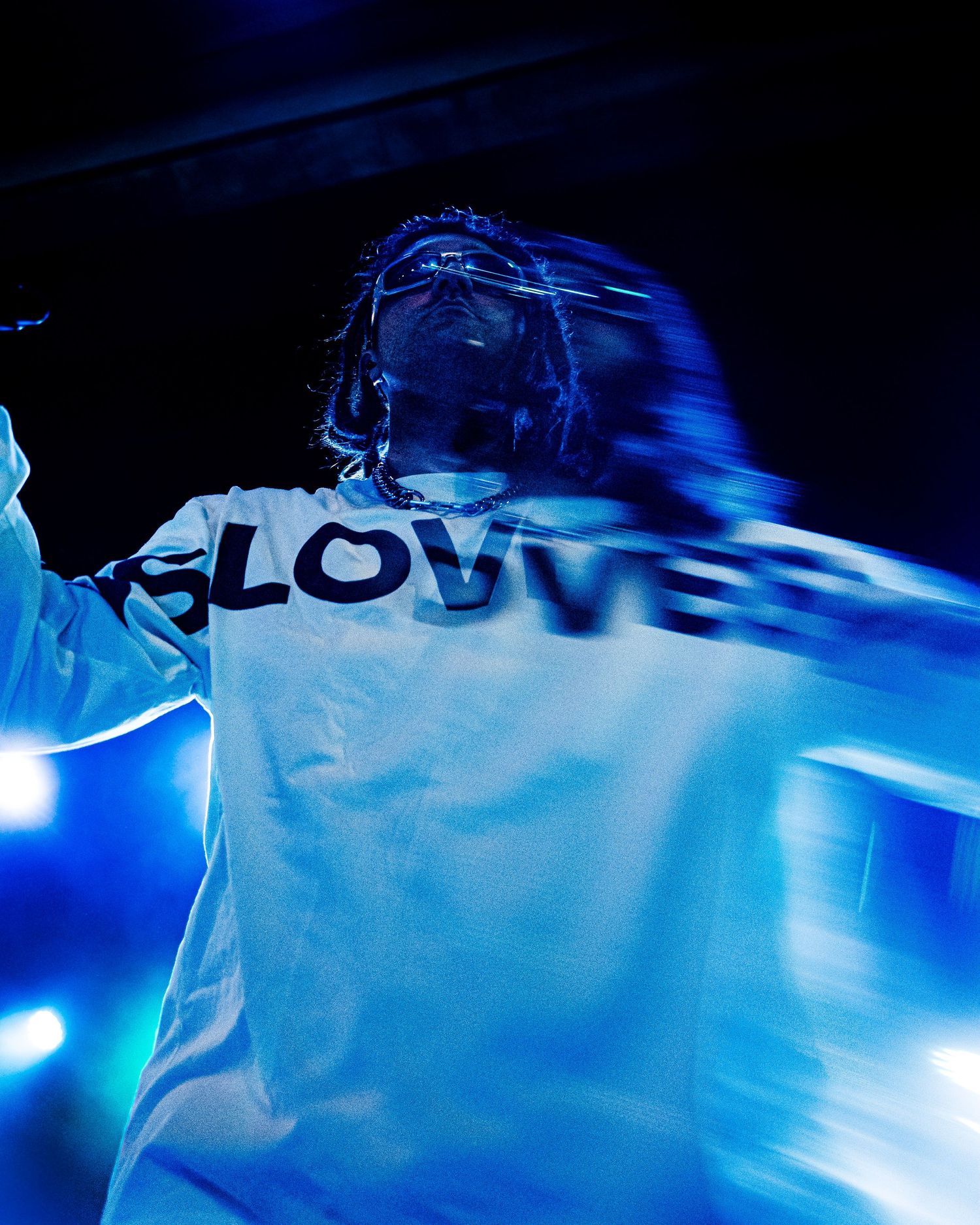 A performer in a white SLOVVE long-sleeve shirt moves dynamically under blue stage lights in a dark venue.
