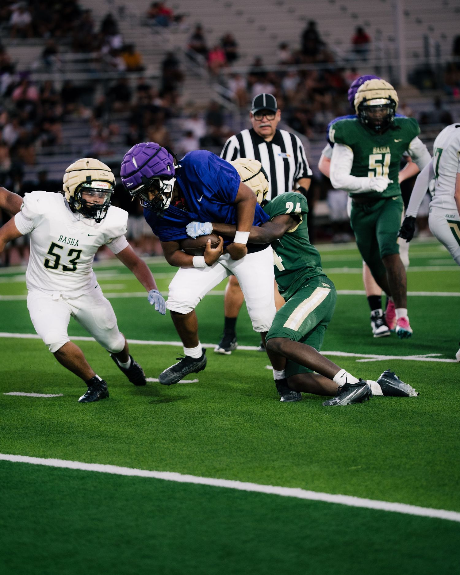 Football player in blue jersey runs with ball while green and gold defenders attempt a tackle on turf field.