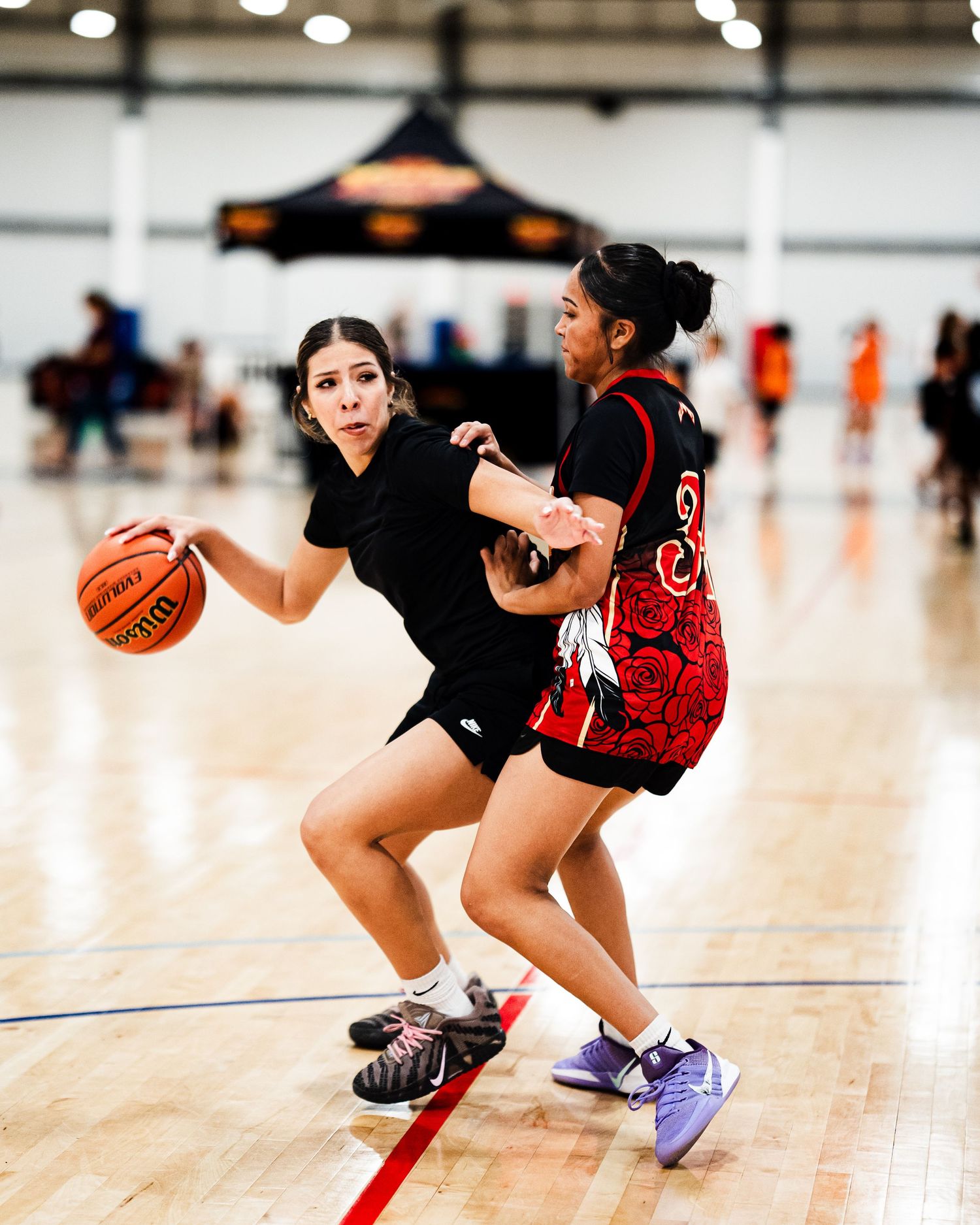 Two female basketball players compete on an indoor court, one dribbling past a defender in a red jersey.
