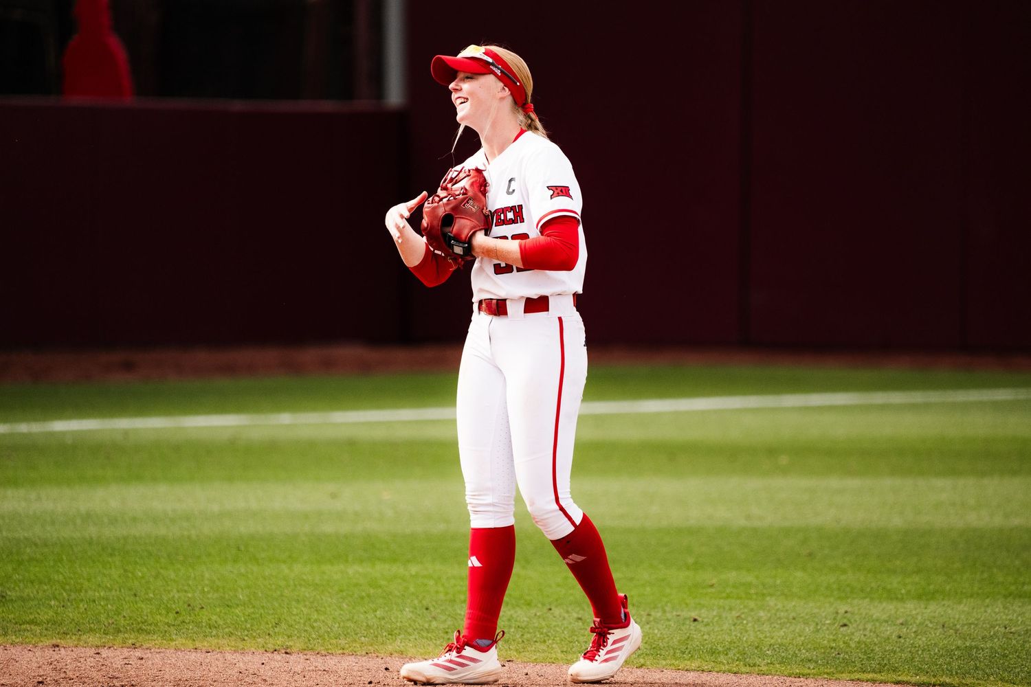 Female softball player in white and red uniform stands on field holding glove, ready for play during a game.