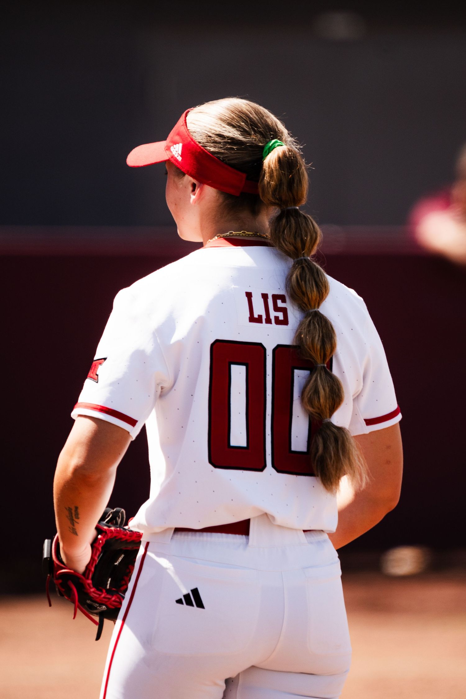 Softball player wearing white jersey with number 00 and name LIS, red cap, holding glove on sunny field.