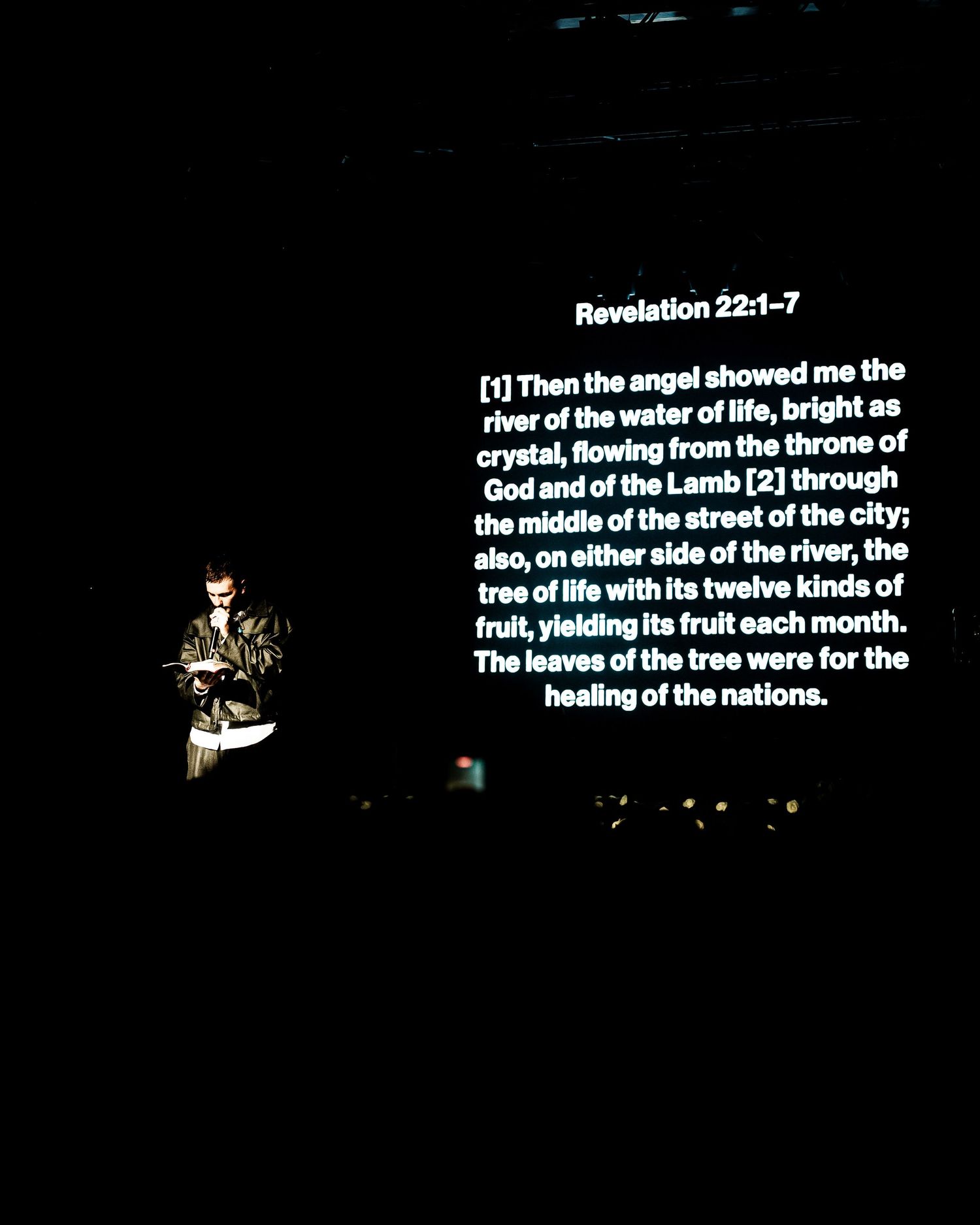 Speaker reads scripture Revelation 22:1-7 on large screen at dimly lit religious event or church service.