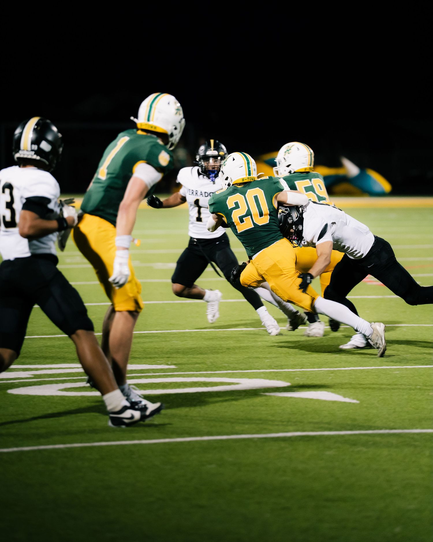 High school football players in green and white uniforms compete during a night game on a well-lit turf field.