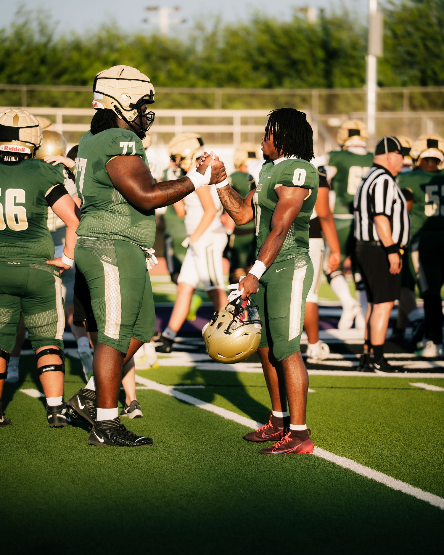 Two football players in green uniforms share a handshake on the sideline during a high school football game.