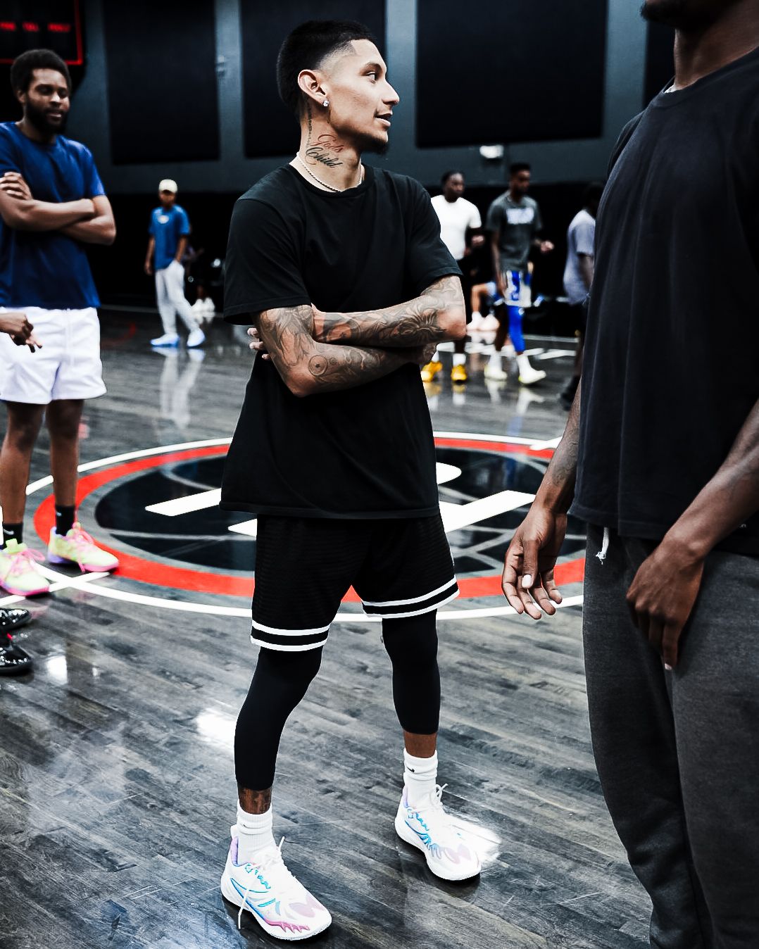 Tattooed basketball player in black outfit and white sneakers stands on indoor court during practice.