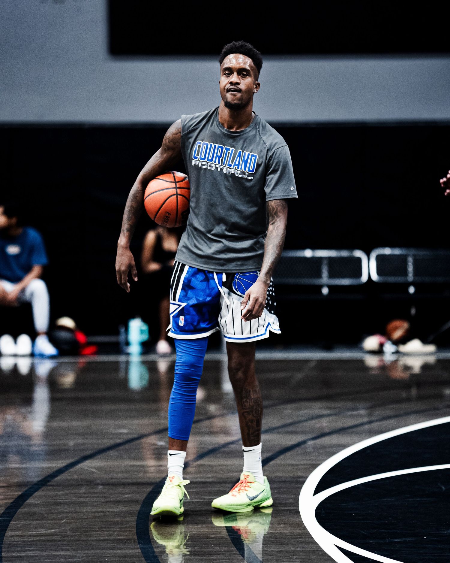 Basketball player in gray Courtland shirt dribbles on a shiny indoor court wearing blue shorts and neon sneakers.