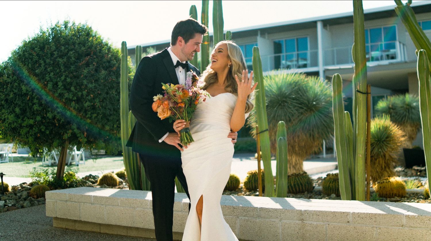 Bride in white gown holds bouquet as groom in tuxedo leans in, surrounded by desert cacti at outdoor wedding venue.