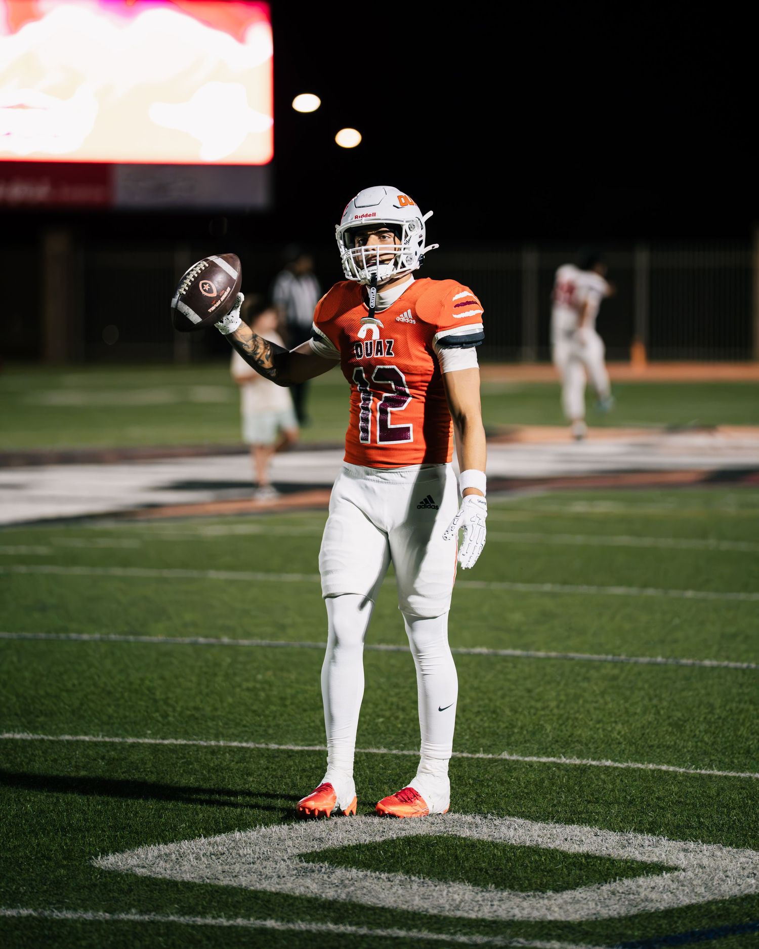 Football player #12 in orange and white uniform holds ball up on field at night game.