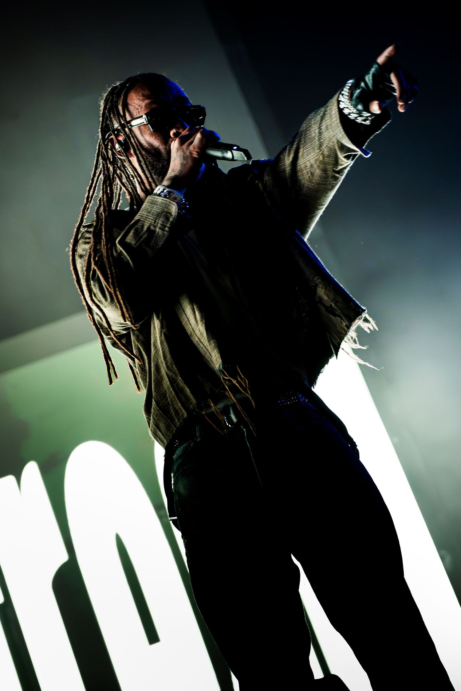 Performer with long dreadlocks and sunglasses sings into a microphone while pointing upward on a dimly lit concert stage.