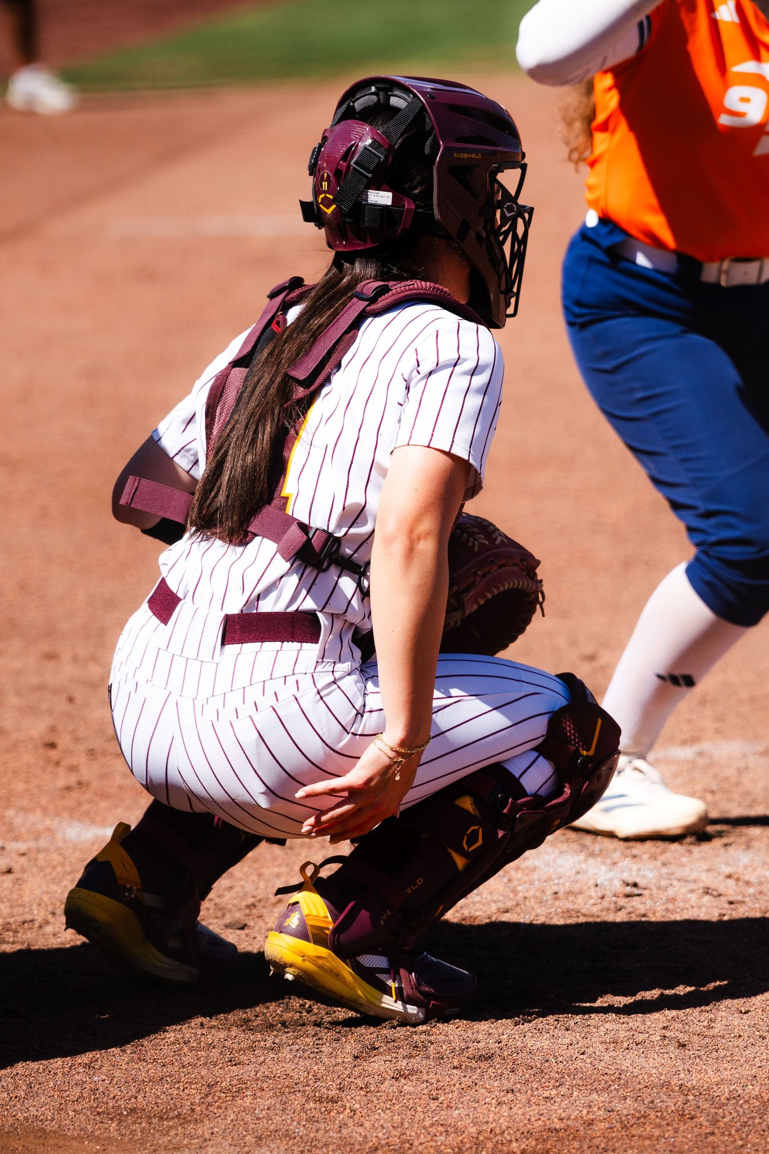 Female softball catcher in maroon pinstripe uniform crouches in position during a college softball game.
