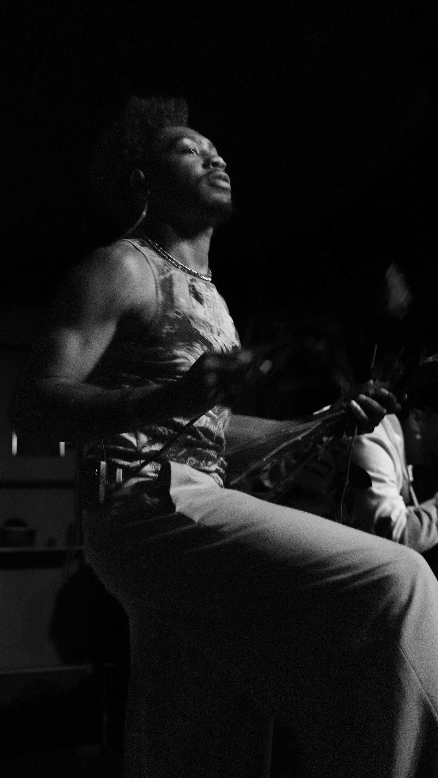 Black and white close-up of a male performer with natural hair sitting and holding a microphone on stage.