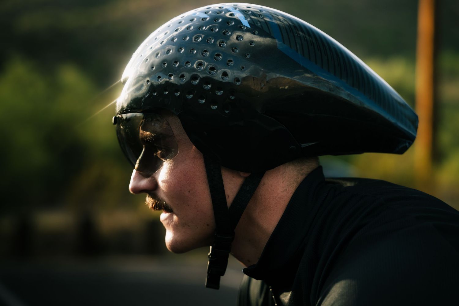 Side profile of male cyclist with mustache wearing black aero helmet and sunglasses in golden light.