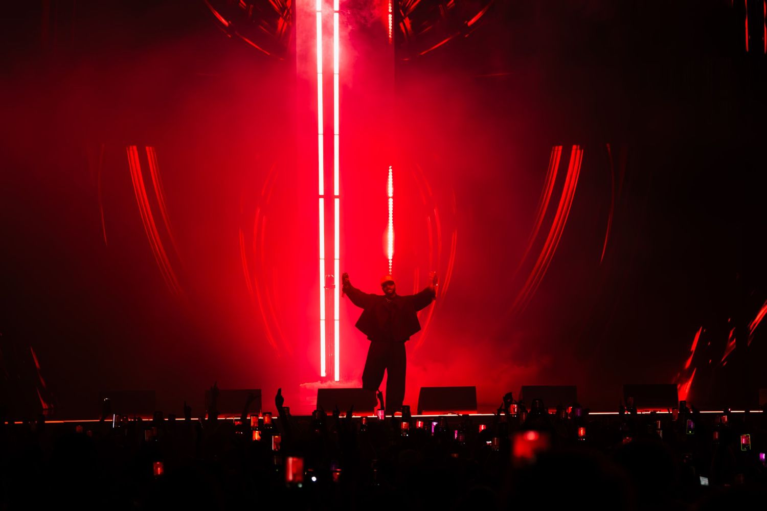 Silhouetted performer with arms raised on a dark stage bathed in vivid red light beams, facing a crowd at a live concert.