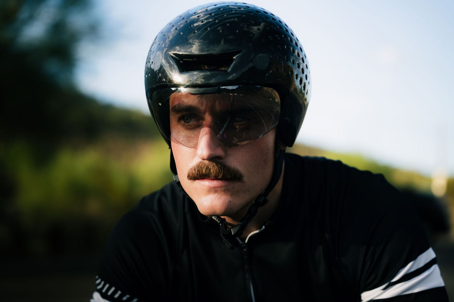 A cyclist wearing a black helmet and sunglasses with a mustache poses outdoors in cycling gear.