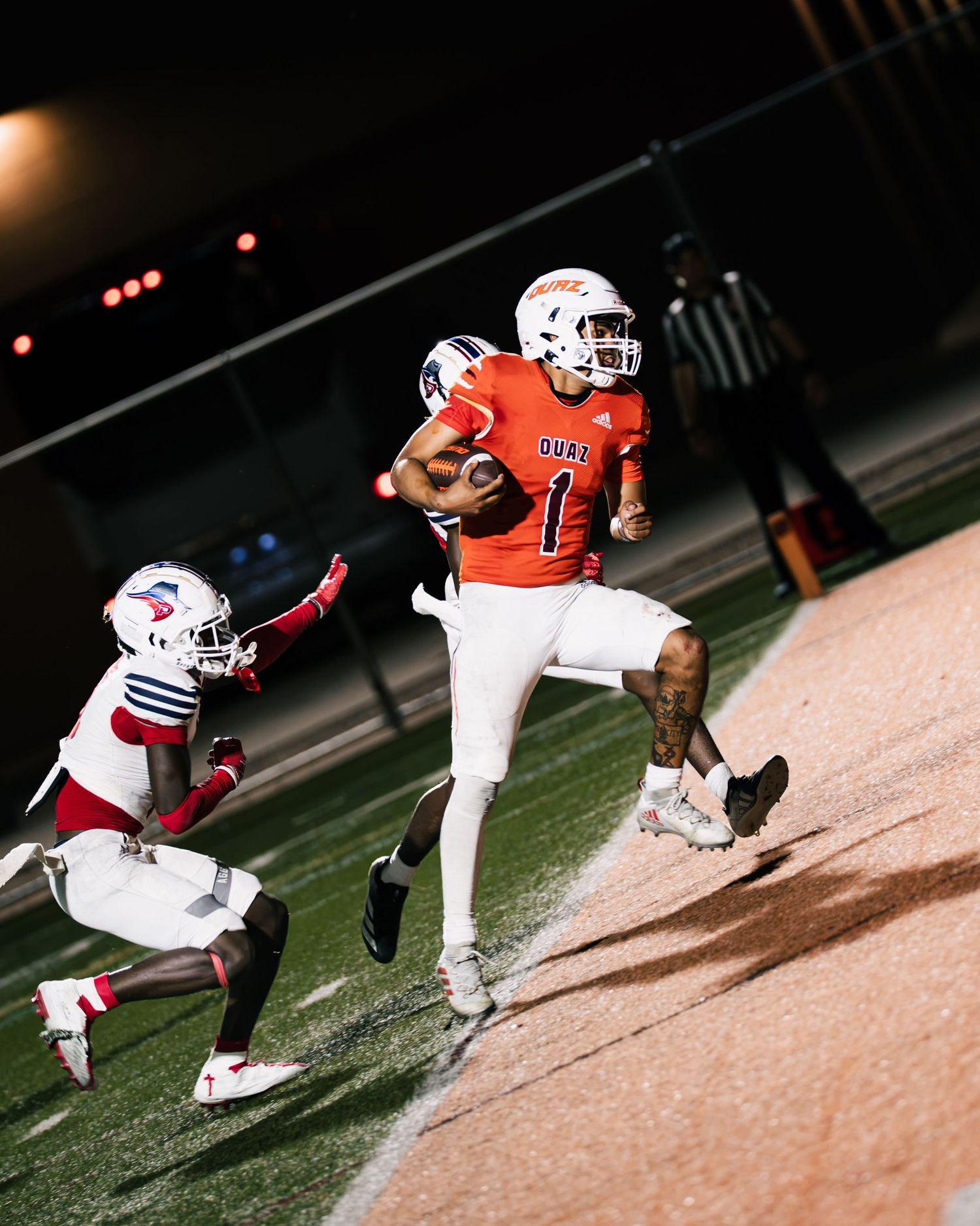 Football player in orange jersey #1 sprints along sideline evading defenders during a night game.