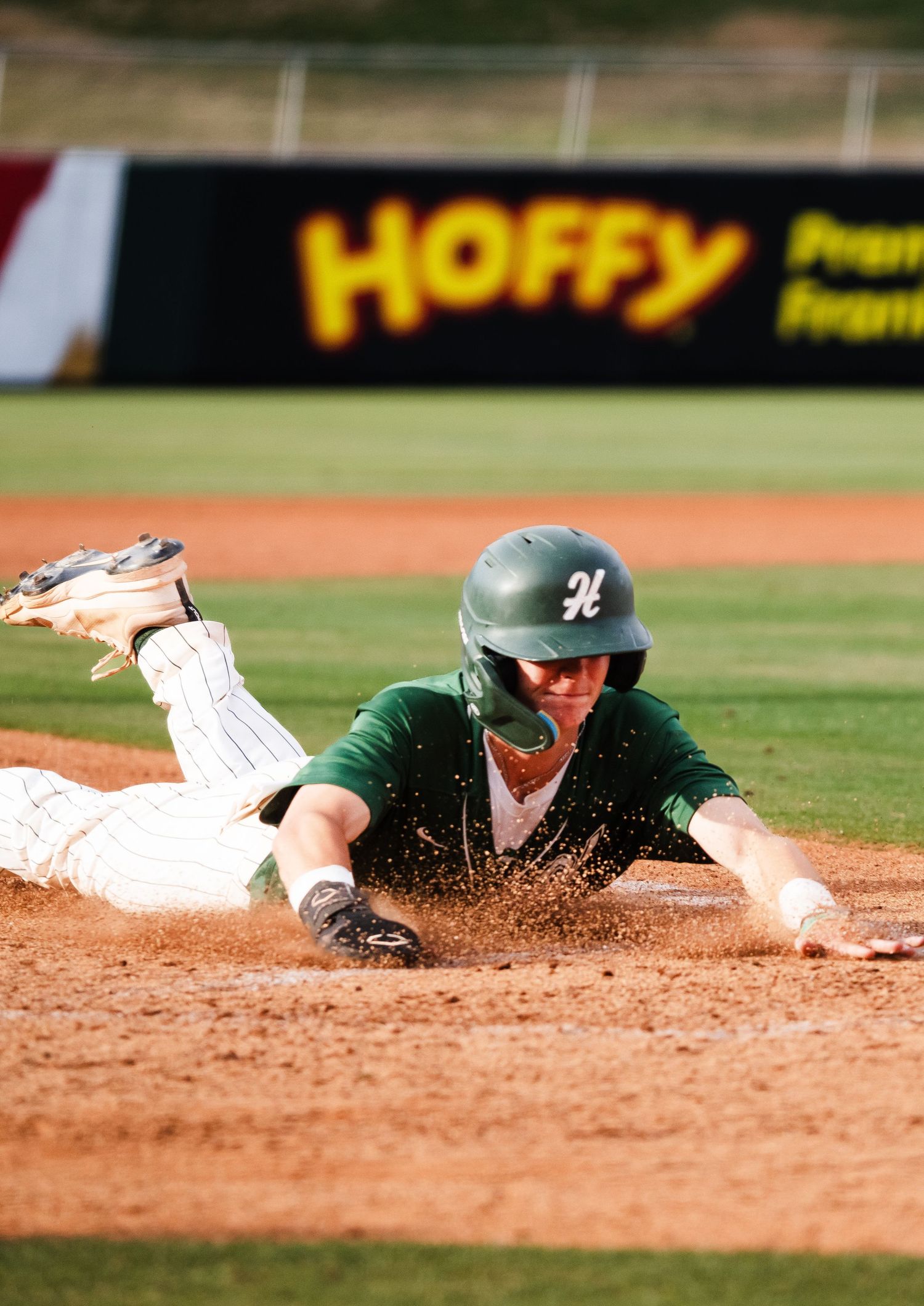 Baseball player in green uniform diving headfirst into base, kicking up dirt, with Hoffy advertisement in background.