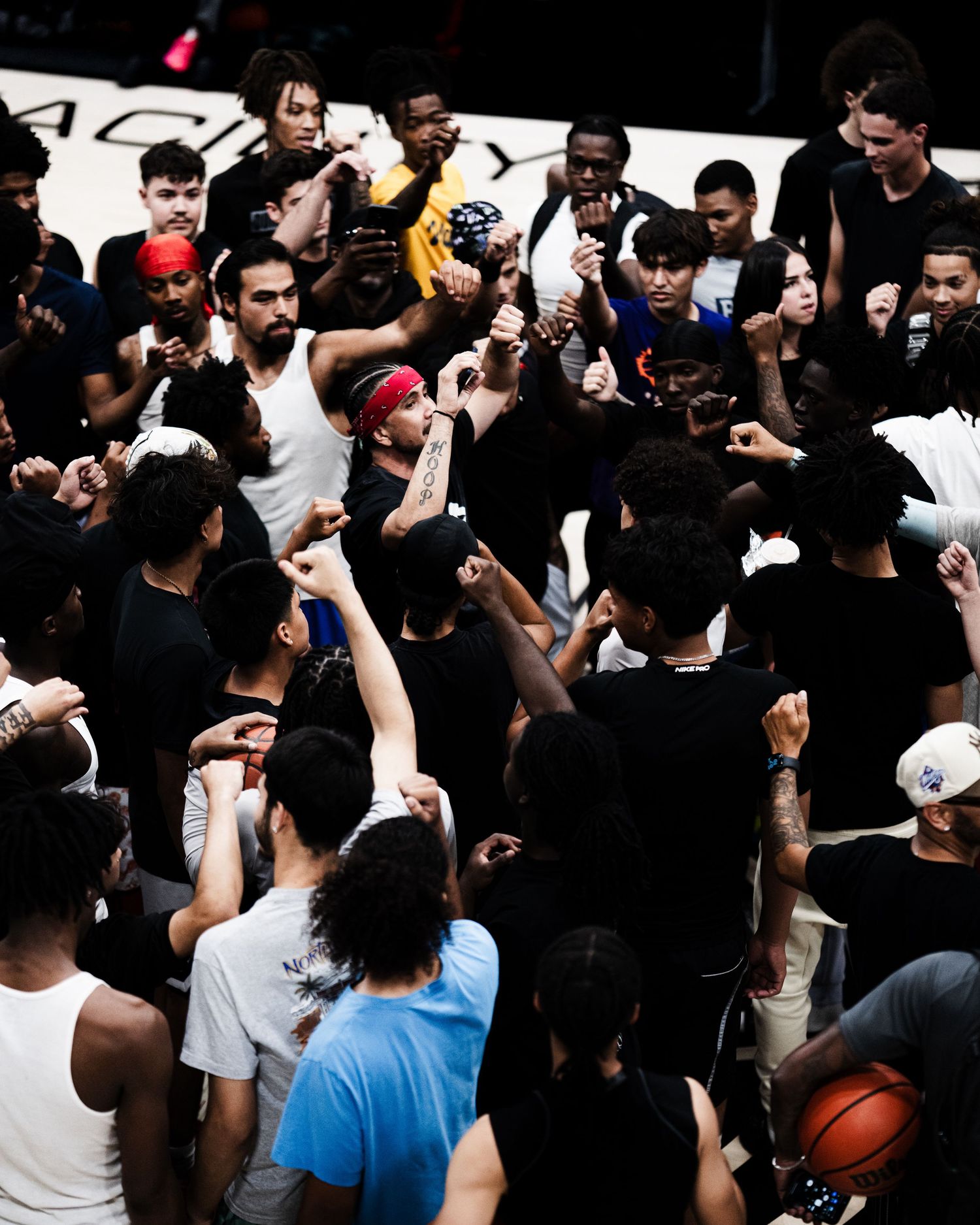 A diverse group of basketball players huddle together raising fists on an indoor court.