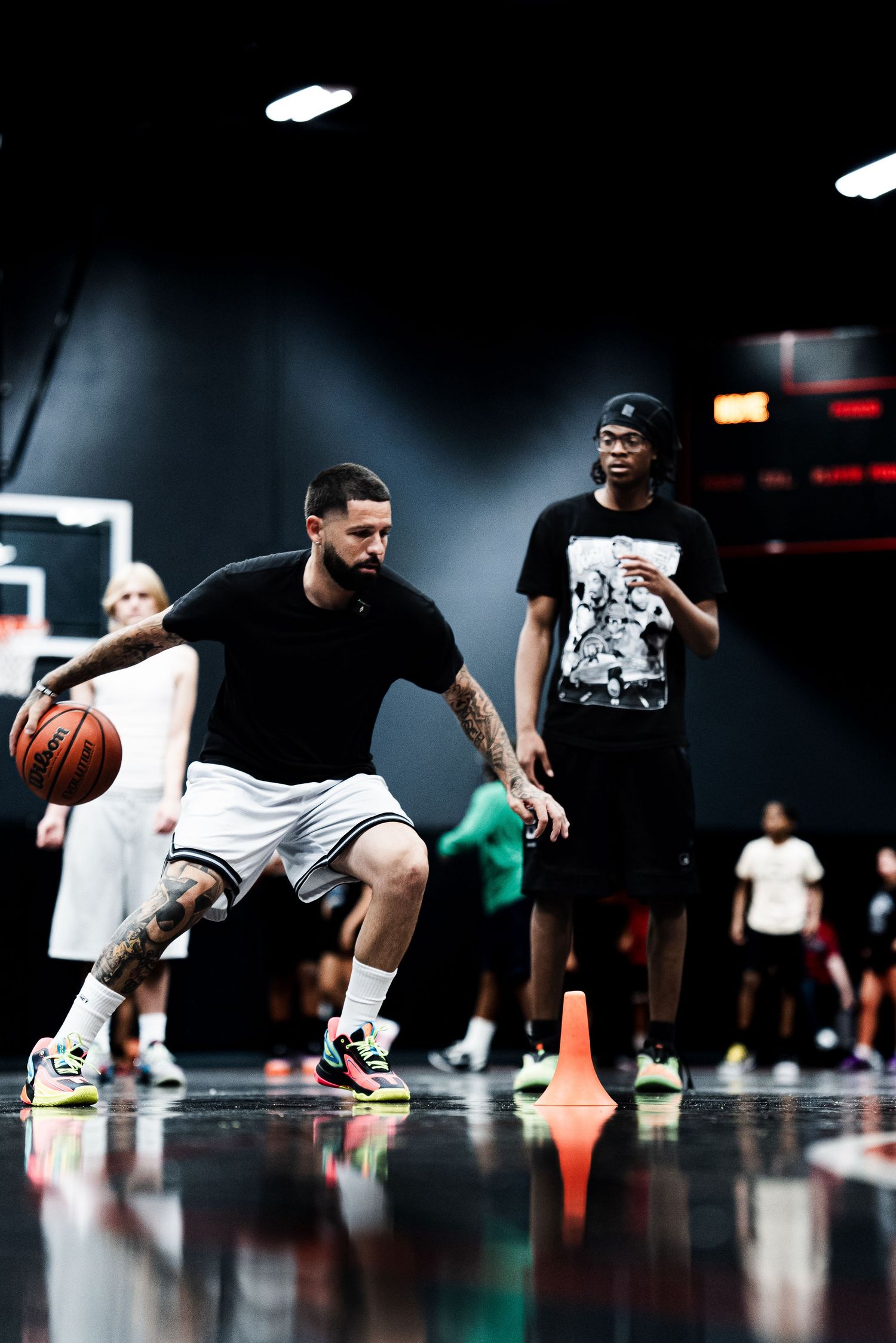 A tattooed player dribbles a basketball past a cone during an indoor training session.