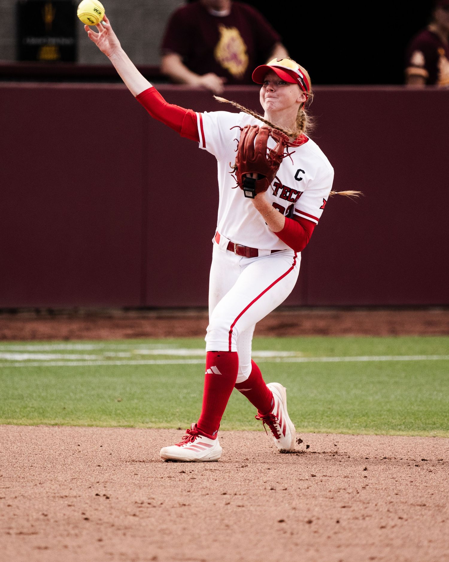 Female softball player in red and white uniform throwing ball during a college game.