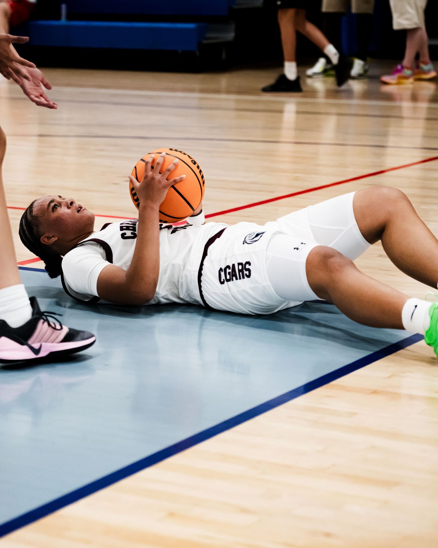 A female basketball player in a white Cougars uniform lies on the court holding a basketball during a game.