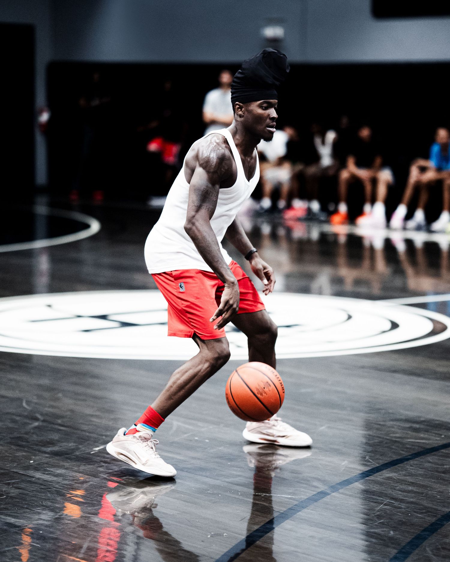 Tattooed basketball player dribbling ball on indoor court wearing white jersey and red shorts.