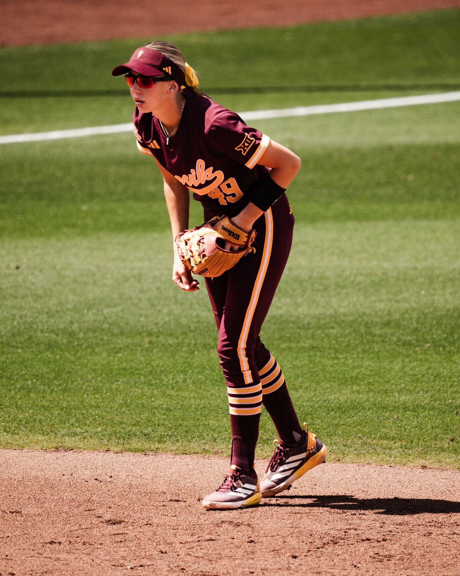 Female softball player in maroon and gold uniform crouches in fielding position on dirt infield during game.