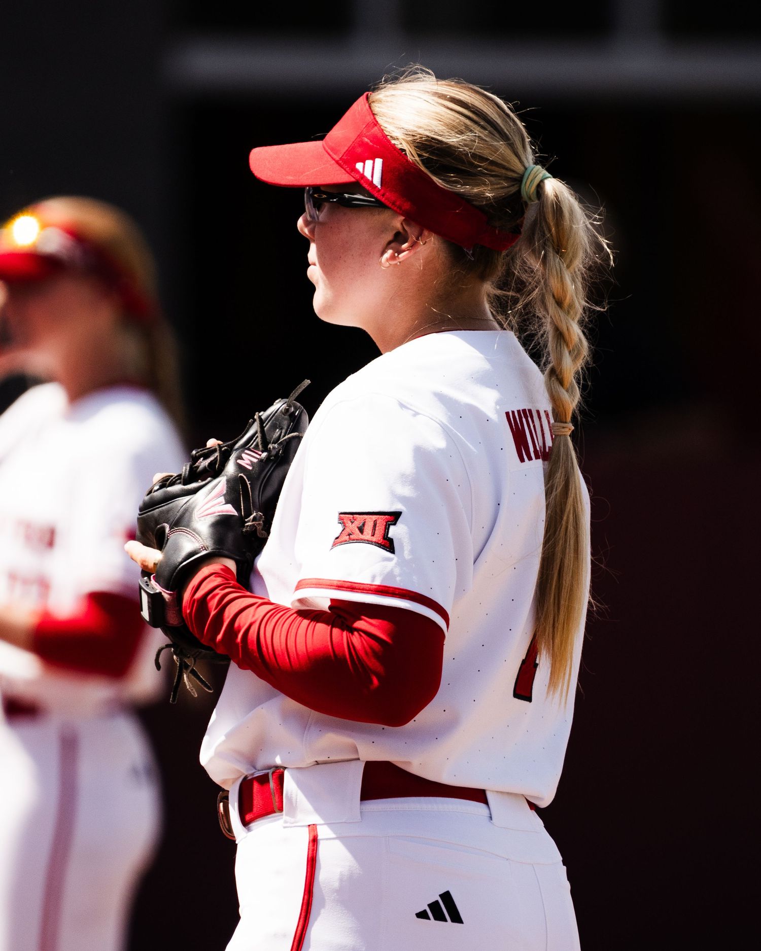 Female softball player in red and white Big 12 uniform holding glove on sunny game day.