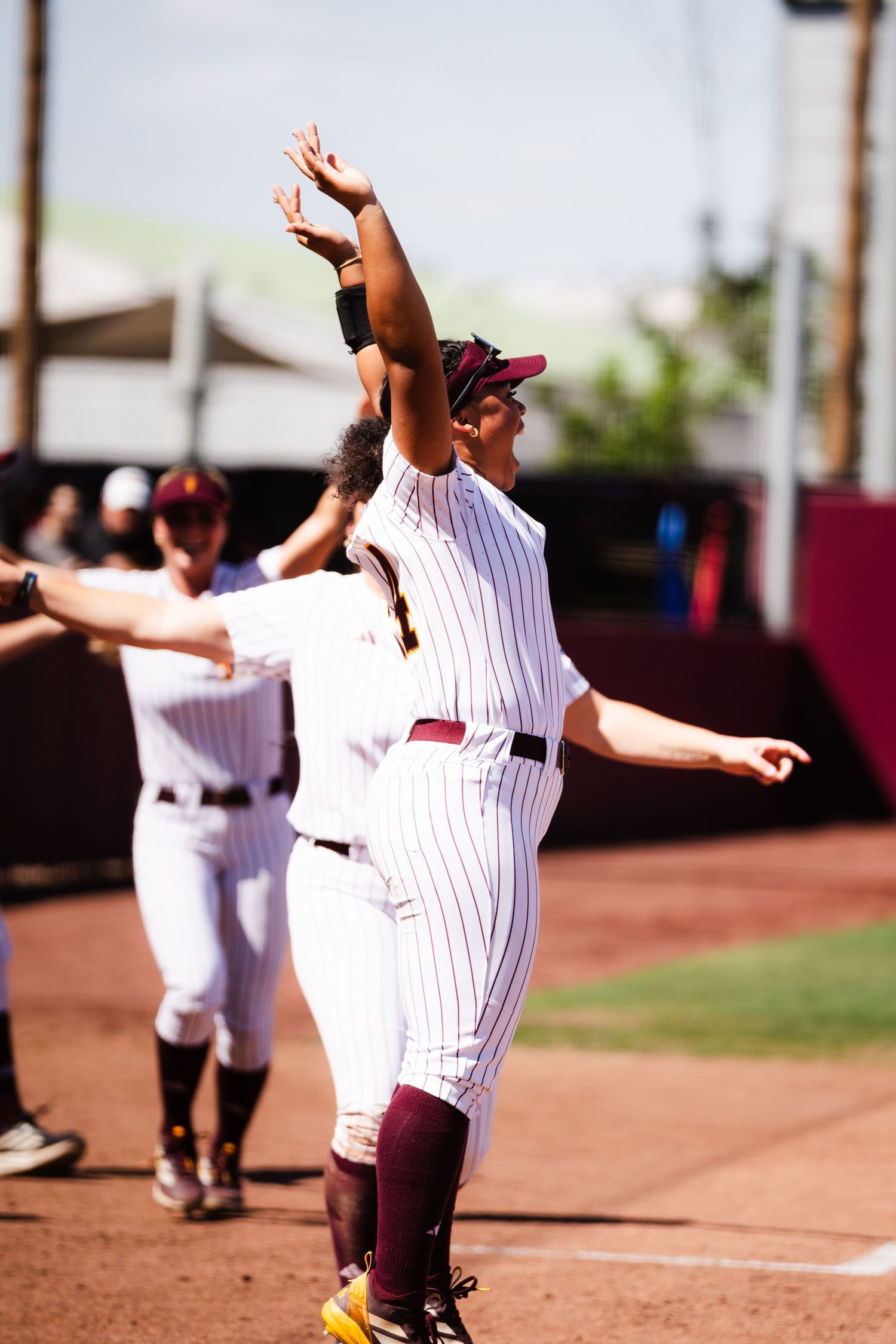 Female college softball player in maroon pinstripe uniform celebrates with arms raised during a game.