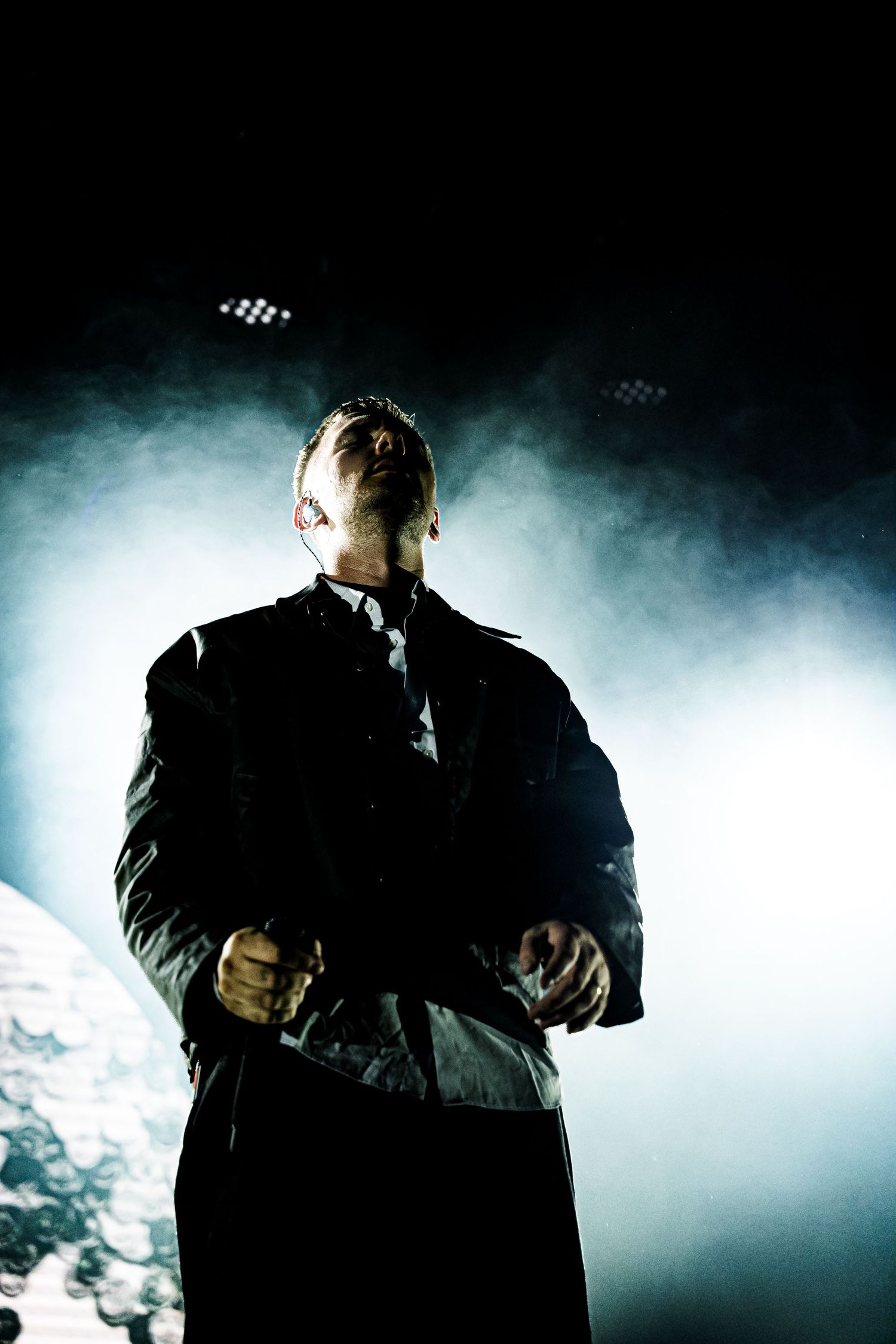 A dramatic backlit performer stands on stage surrounded by smoke and spotlight during a live concert.