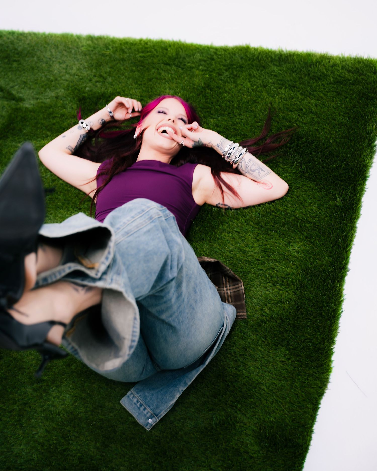 Woman with red hair lying on green turf, smiling and making peace signs, wearing purple top and jeans.