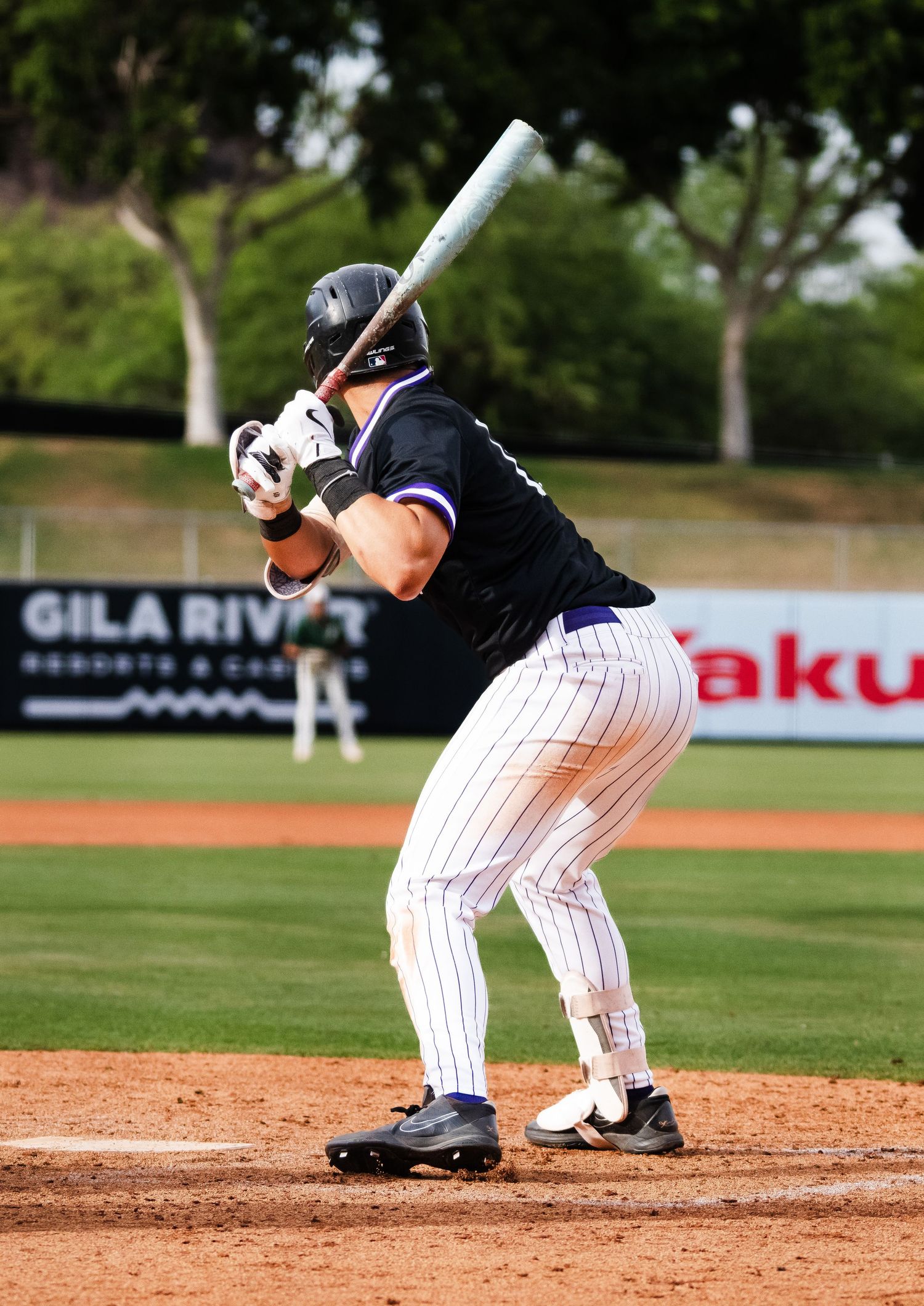 College baseball batter in black pinstripe uniform preparing to swing at pitch near Gila River advertisement.