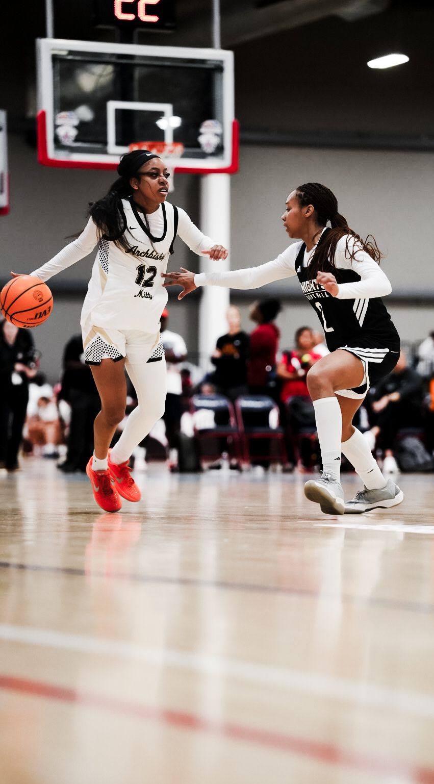 Female basketball player #12 in white Archbishop jersey drives past defender during indoor high school game.