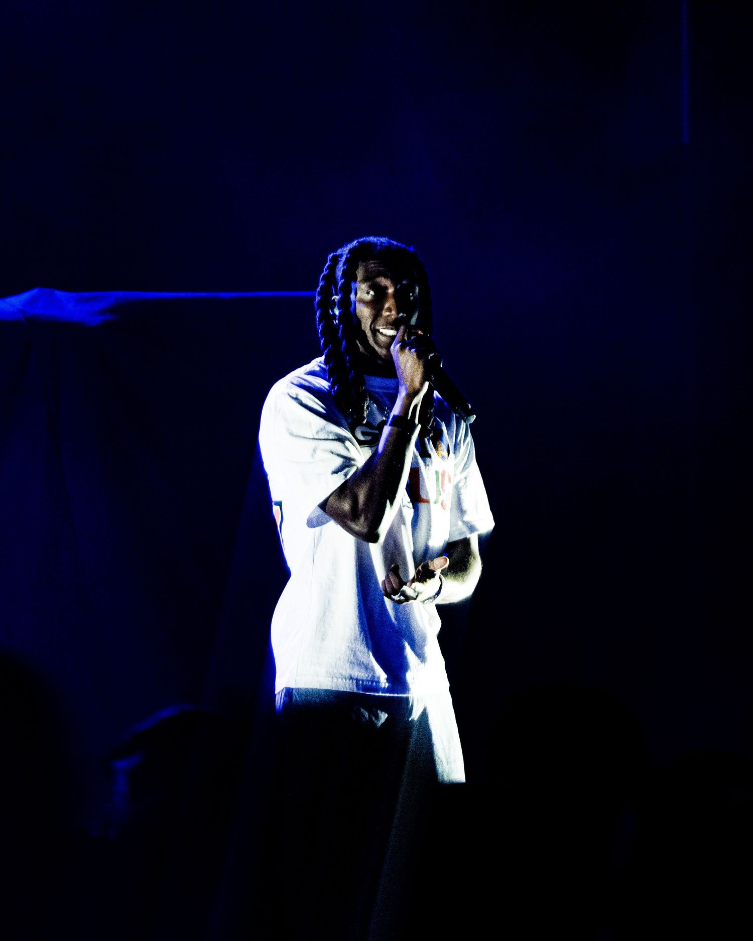 A performer in white clothing holds a microphone on a dark, blue-lit stage during a live concert.