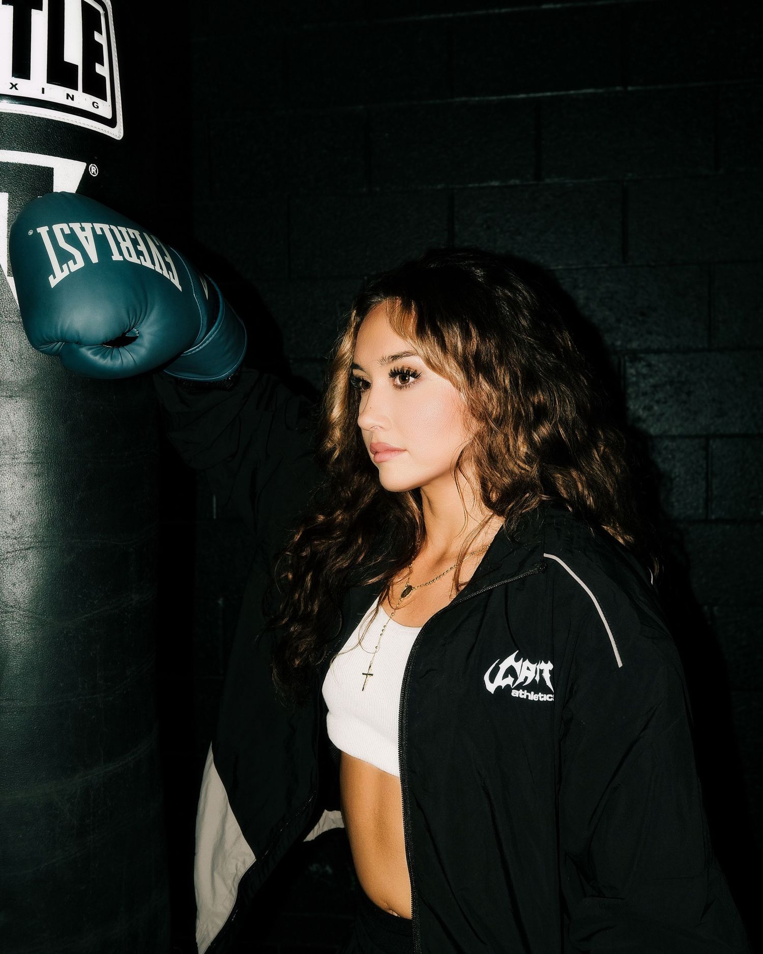 Woman with long wavy hair in a black jacket and white crop top rests a teal Everlast boxing glove on a heavy punching bag in a dark gym.