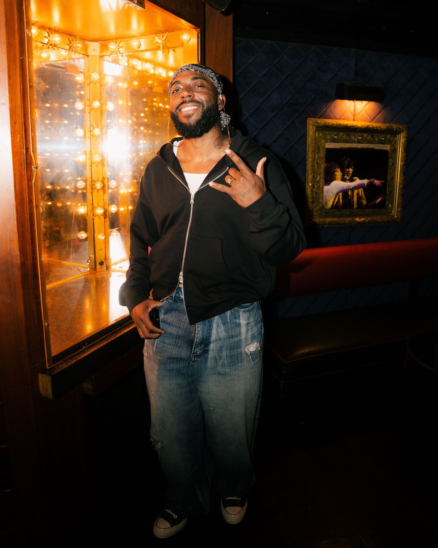 A smiling man in a black hoodie and jeans poses with a hand gesture in a warmly lit venue with decorative framed art.