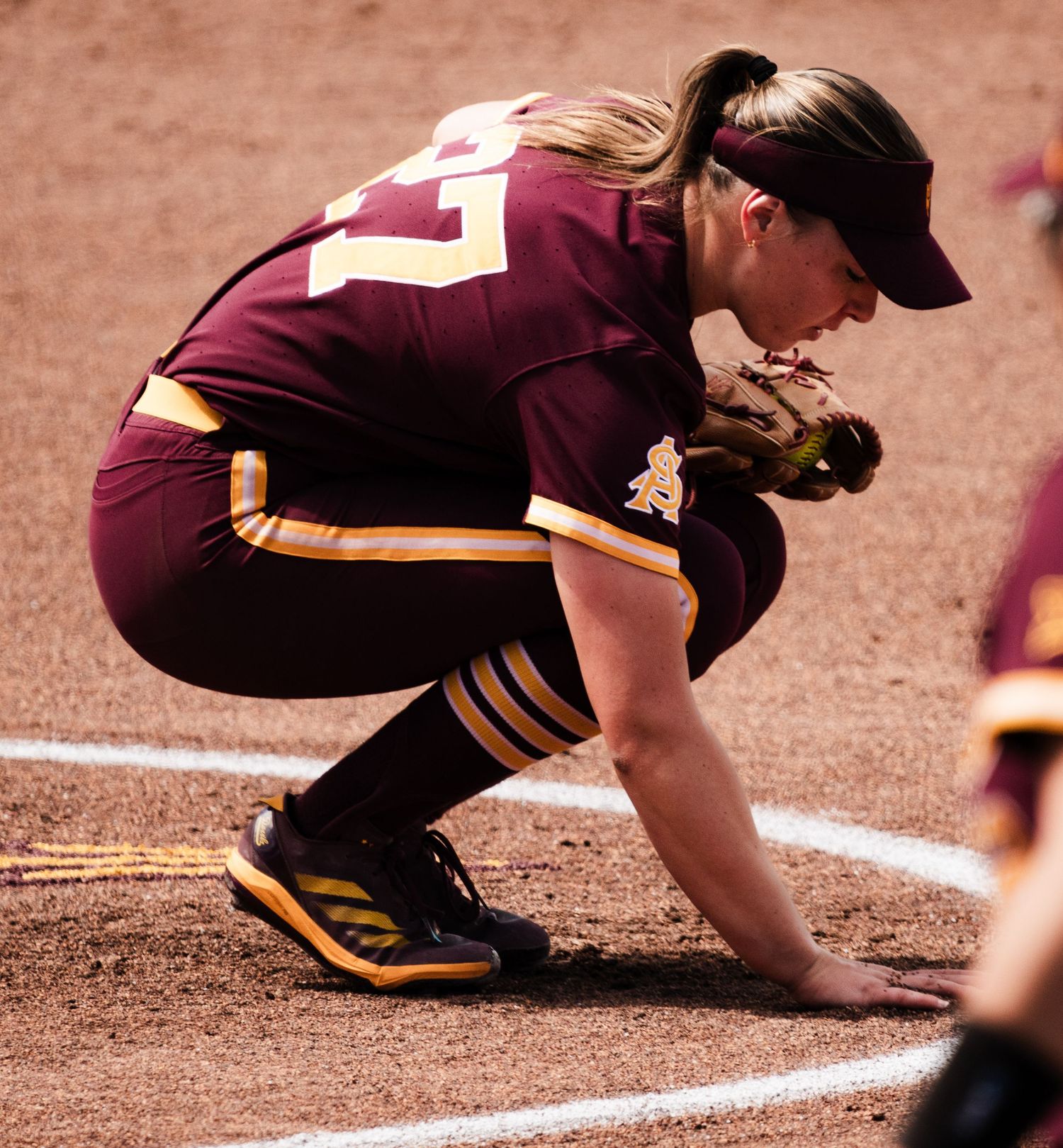 Female softball player in maroon uniform crouching at base during game, wearing cap and fielding glove.