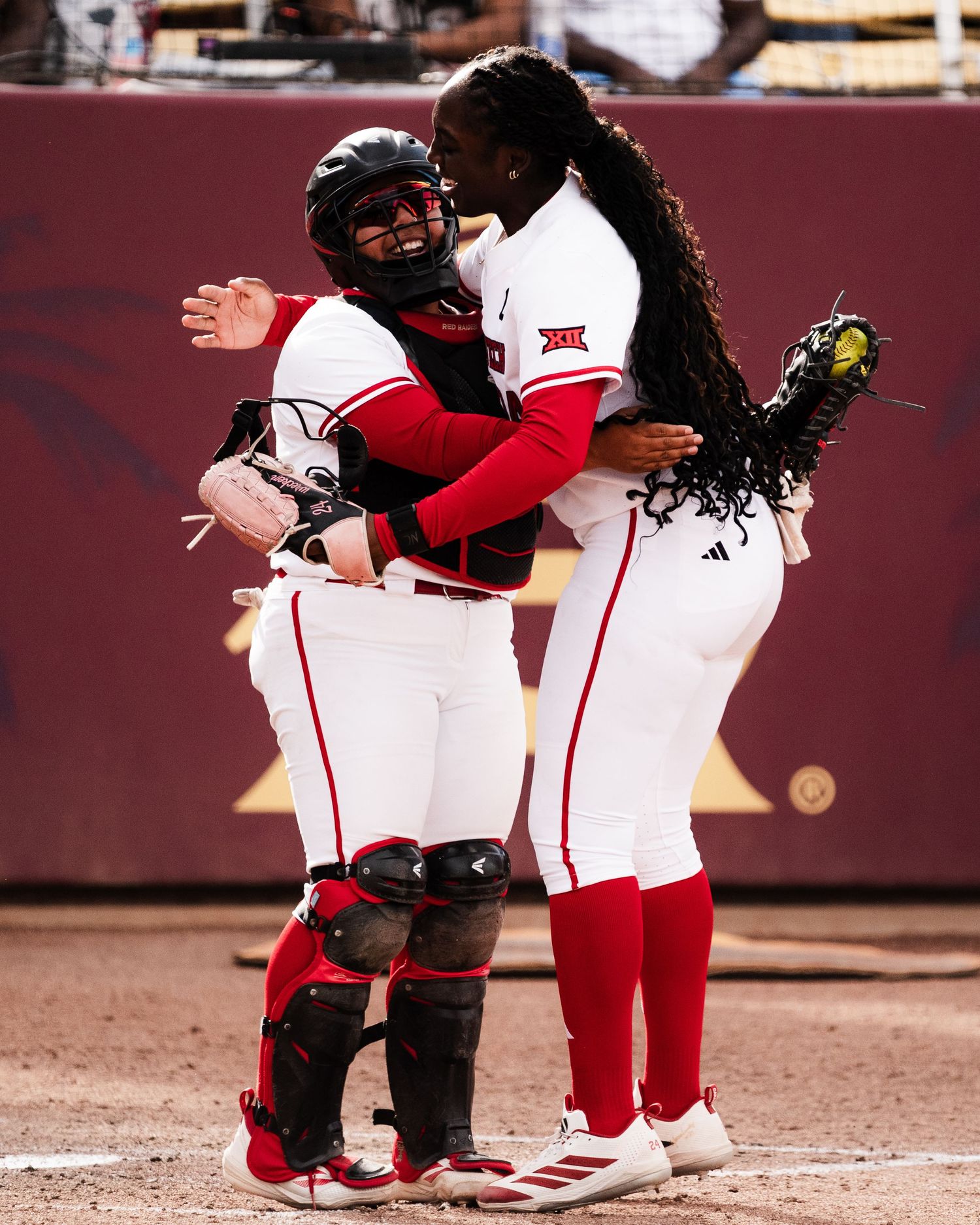 Two college softball players in red and white uniforms share a celebratory hug on the field during a game.