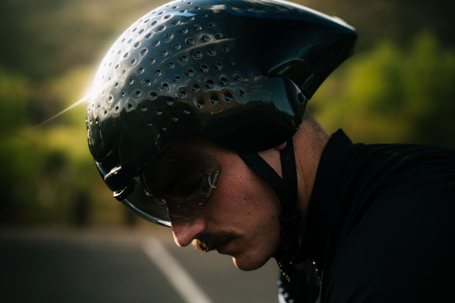 Cyclist wearing a sleek black aerodynamic helmet with ventilation holes, shot in profile against a sunlit mountain road.
