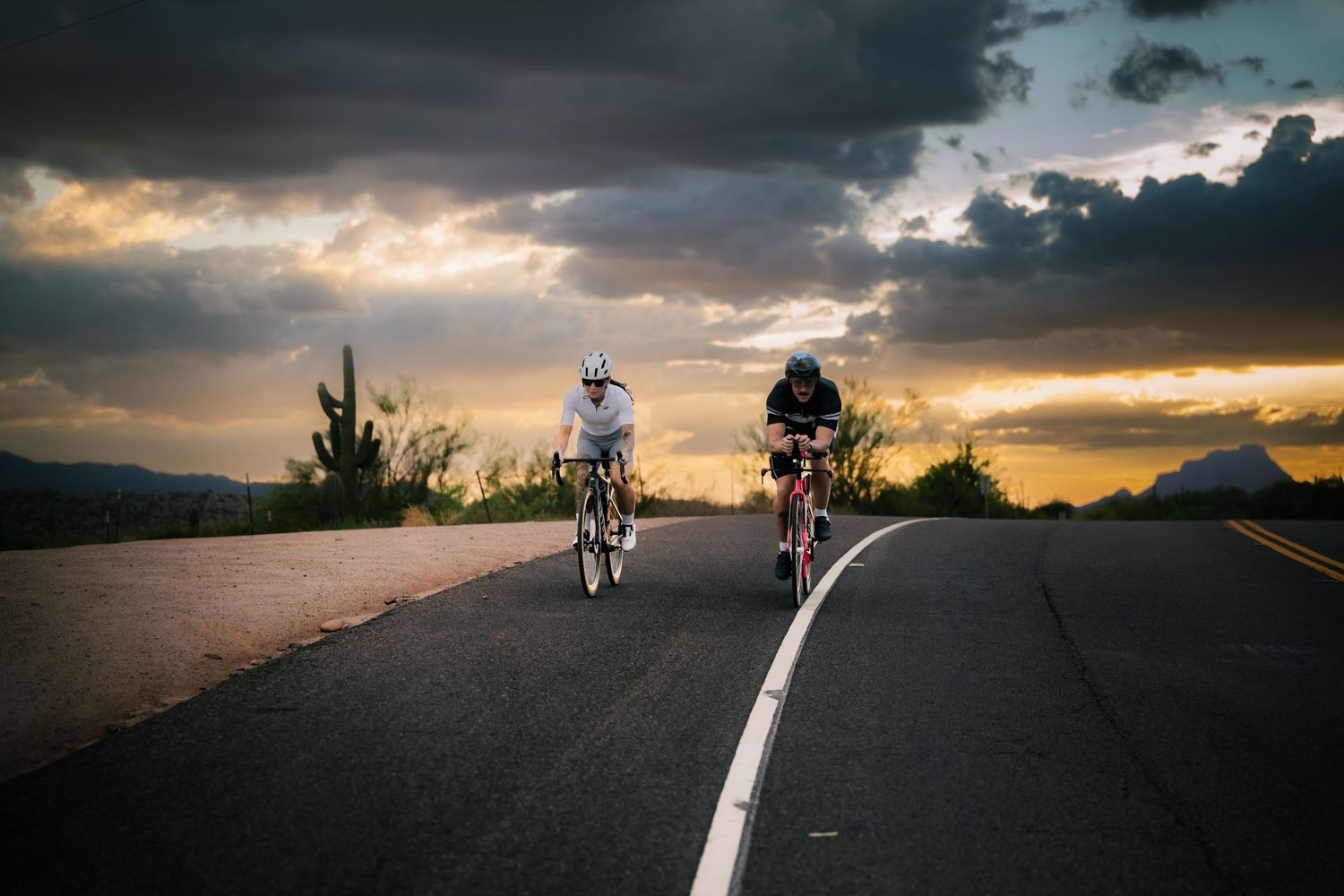 Two cyclists ride on a winding road at sunset with dramatic storm clouds and mountains in the background.