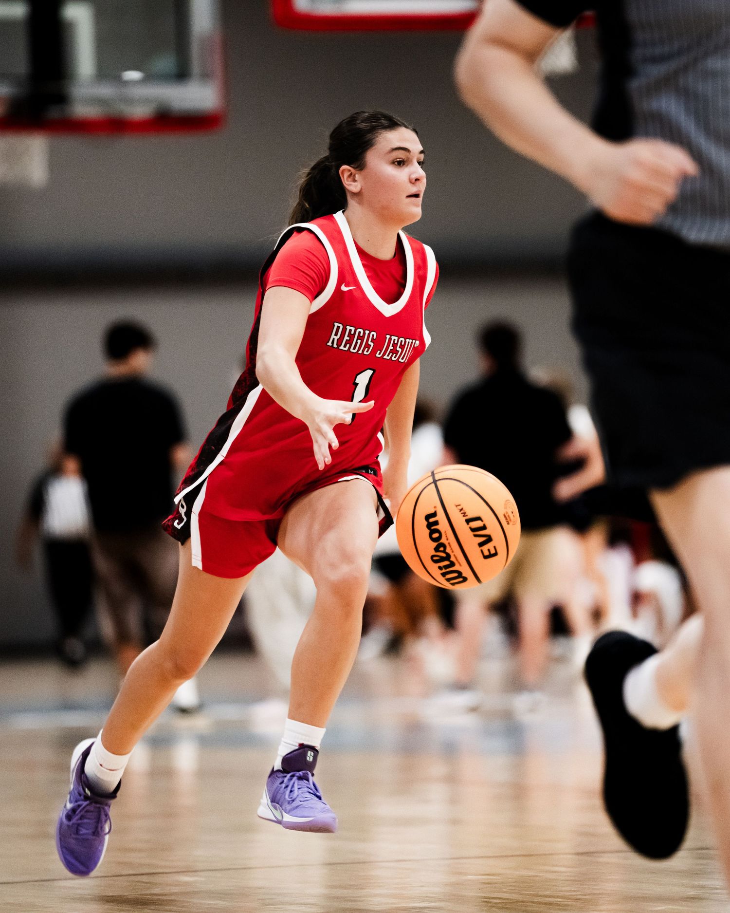 A female basketball player in a red Regis Jesuit number 1 jersey dribbles the ball down the court.