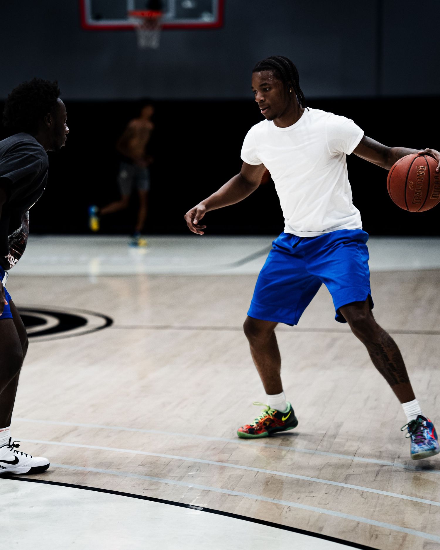 Two basketball players practice dribbling drills on an indoor court, one in a white tee and blue shorts holding the ball.