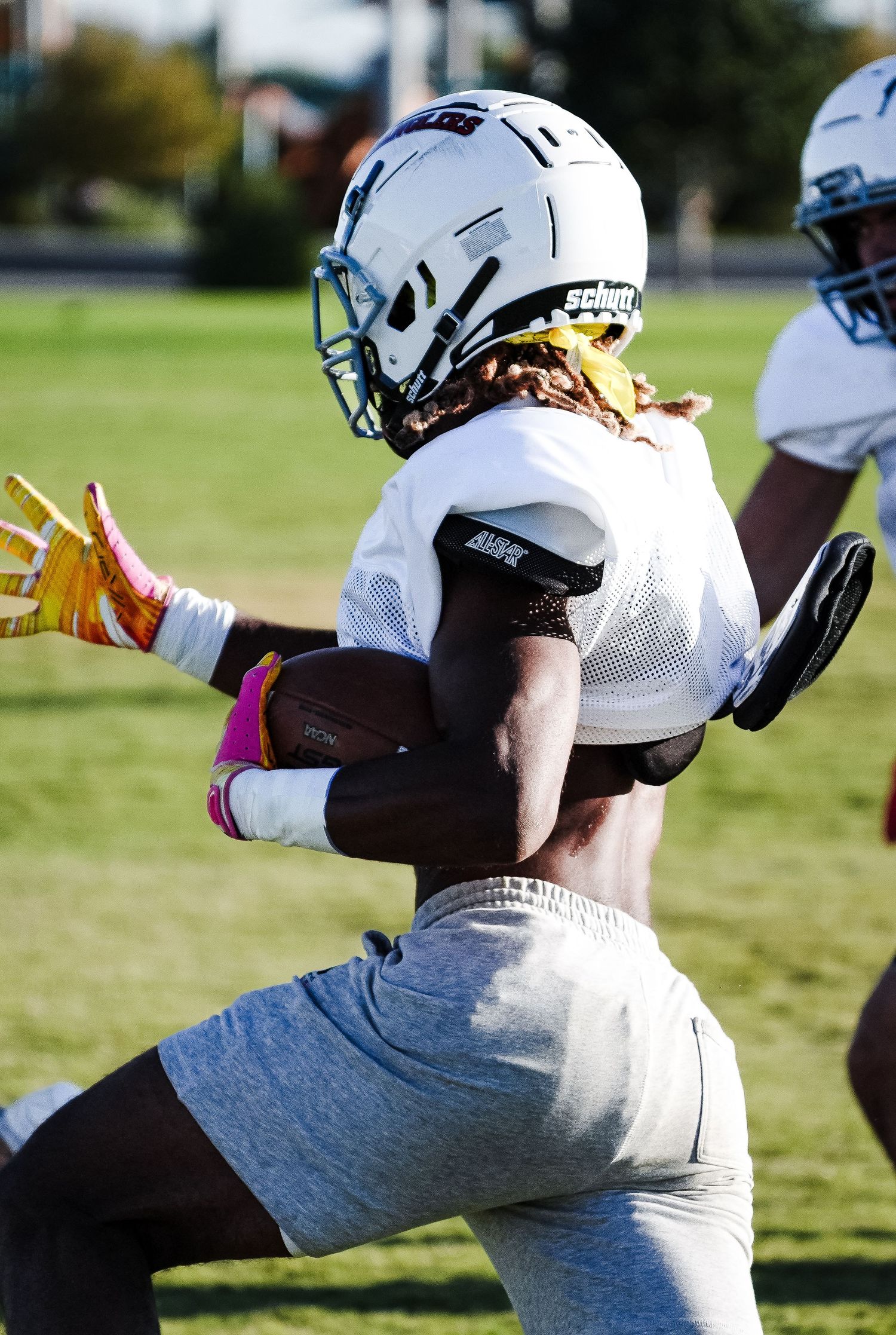 Football player in white helmet and jersey carries ball during outdoor practice drill on green field.